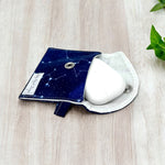 Load image into Gallery viewer, Open navy blue constellation print AirPods pouch with white earbuds case inside, resting on light wood surface with green leaves nearby
