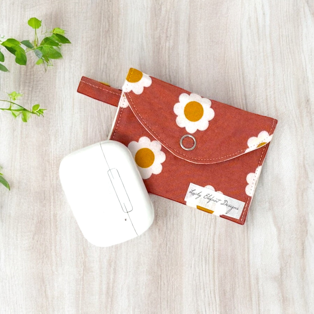 Rust-colored daisy print AirPods pouch with snap closure and white earbuds case on light wood background