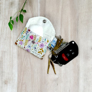 Open floral fabric pouch with snap closure holding AirPods, attached to a keyring with car keys and gold keys on light wooden surface