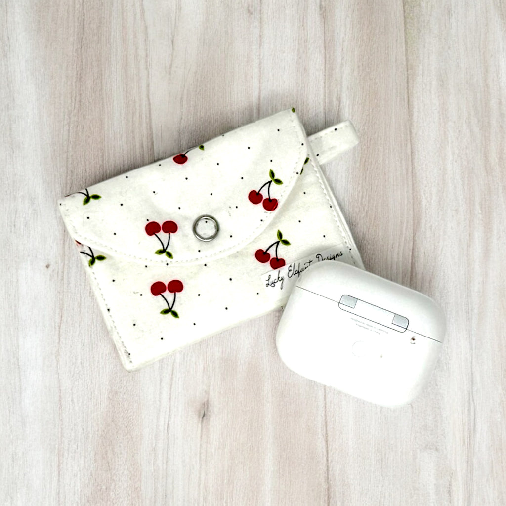 White fabric AirPods pouch with red cherry print and snap closure next to wireless earbuds case on wood surface