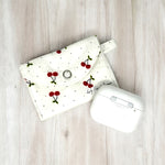 Load image into Gallery viewer, White fabric AirPods pouch with red cherry print and snap closure next to wireless earbuds case on wood surface
