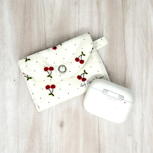 White fabric AirPods pouch with red cherry print and snap closure next to wireless earbuds case on wood surface