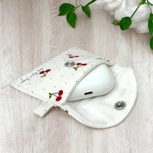 Open cherry print fabric AirPods pouch showing wireless earbuds case inside on light wood surface with green leaves and cotton