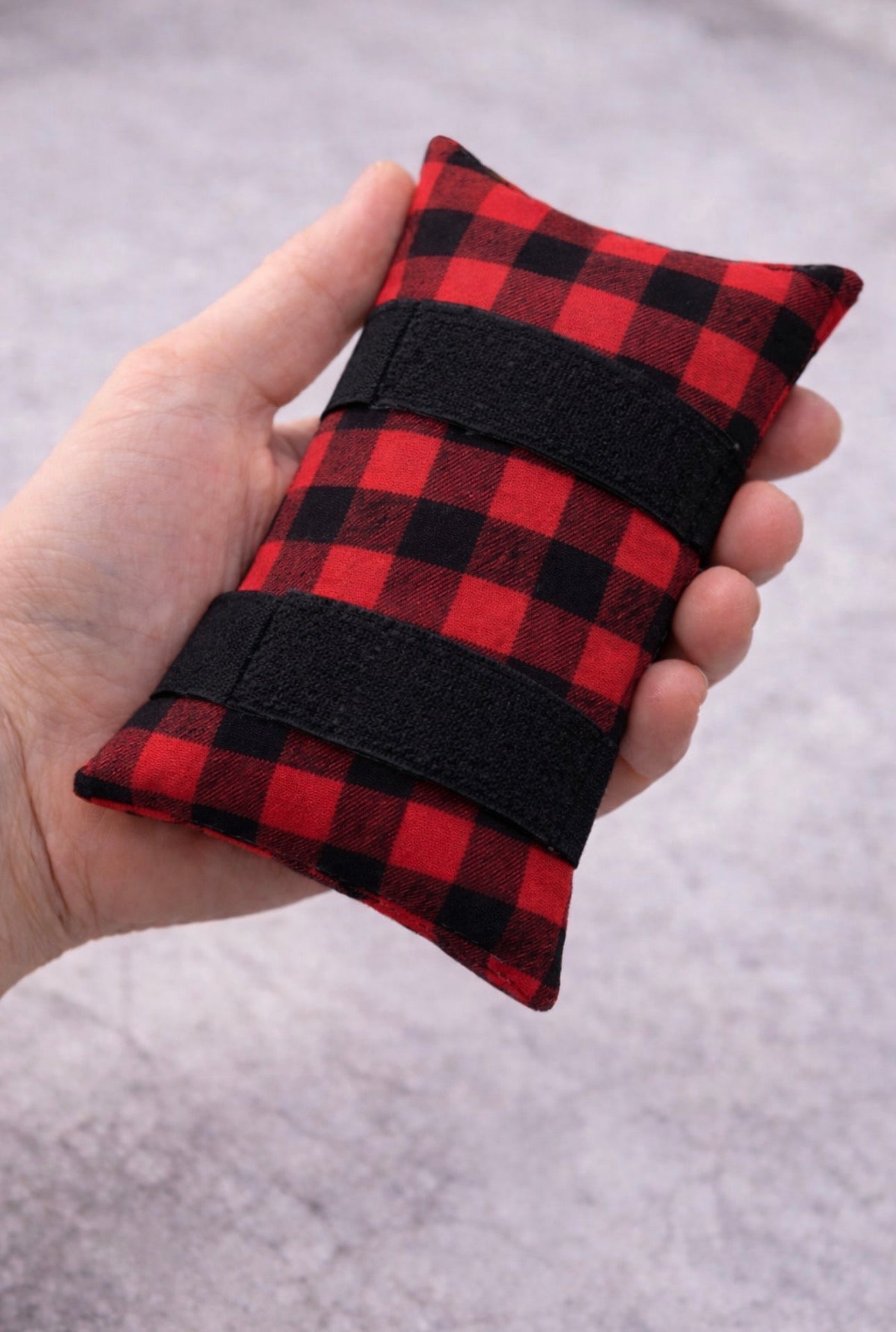 Hand holding red and black plaid chemo port pillow with soft padding and Velcro straps designed to reduce seatbelt pressure on chest port area